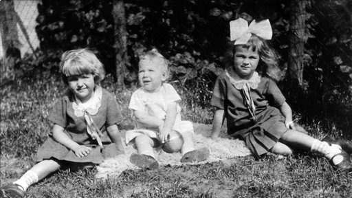 Eine Schwarz-Weiß-Aufnahme zeigt drei Kinder, die im Graß sitzen