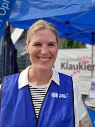 Eine Beamtin der Polizei steht an einem Infostand für Kriminalprävention und lacht in die Kamera.