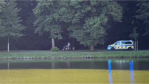 Zwei Menschen sitzen auf einem Motorroller und fliehen vor einem Polizeiwagen.