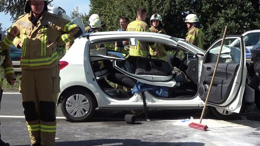 Ein stark demolierter weißer Pkw ist umringt von Einsatzkräften der Feuerwehr.