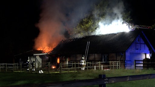 Der Dachstuhl einer Stallung brennt. Feuerwehrleute stehen um das Gebäude.