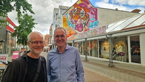 Zwei Männer stehen in einer Fußgängerzone vor Streetart auf einer Hauswand