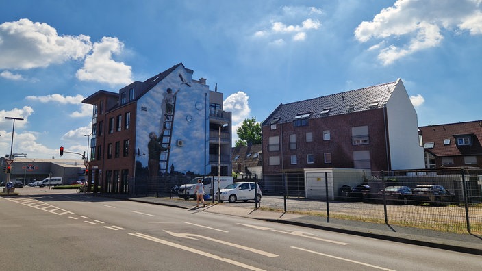 Streetart von Benzi Brofman auf einer Hauswand in Rheine. Ein Mann hält eine Leiter, an der ein Junge hinaufklettert. Im Hintergrund ist die Innenstadt von Rheine zu sehen.
