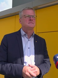 Ein Mann mit Brille steht vor einer Poststation und gibt ein Interview