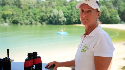 Eine Frau steht neben einem Tisch, auf dem ein Fernglas steht. Im Hintergrund ist ein See zu sehen. 