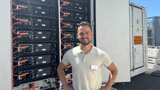 Ein Mann mit braunen Haaren und Bart steht vor einem geöffneten Großbatteriespeicher.