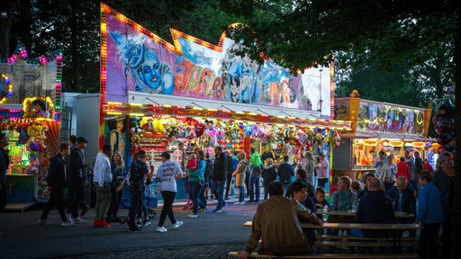 Eine Spielbude auf der Kirmes in Warendorf. Im Vordergrund sitzen Menschen an Bierzeltgarnituren.