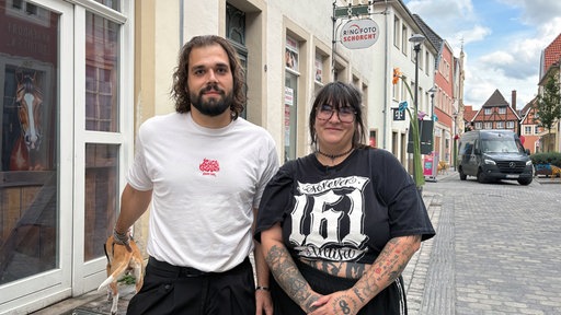 Das junge Paar Kevin Klein und Julia Westerwlad schauen in die Kamera und Kevin Kleine hält einen Leine für den Hund im Hintergrund.
