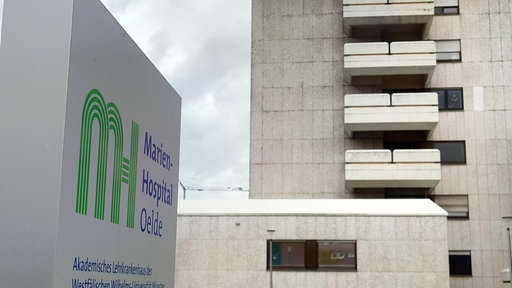 Das Schild des Marien-Hospitals Oelde, im Hintergrund ein graues Gebäude.