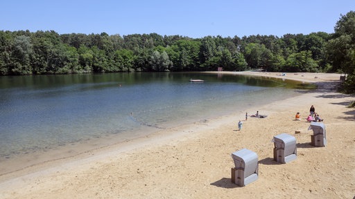 Am Strand des Feldmarksees Sassenberg halten sich Menschen auf. Unweit von ihnen stehen Strandkörbe.
