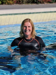 Eine blonde Schwimmlehrerin steht im Schwimmbecken und lächelt in die Kamera.
