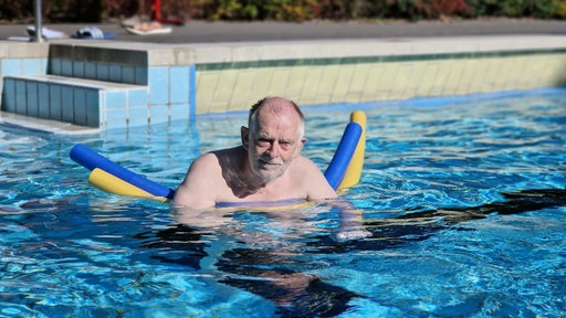 Ein älterer Mann steht mit zwei Schwimmnudeln im Schwimmbecken und blickt in die Kamera.