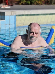 Ein älterer Mann steht mit zwei Schwimmnudeln im Schwimmbecken und blickt in die Kamera.