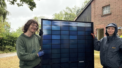 Ein Mann und eine Frau halten ein Solarpanel für ein Dach fest und lächeln in die Kamera.