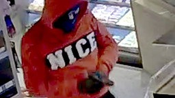 Vermummter Mann mit einem orange-roten Pullover und einer Pistole.