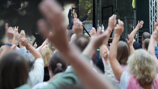 Auf der Bühne wird Stimmung gemacht, während im Publikum Menschen in die Hände klatschen und singen.