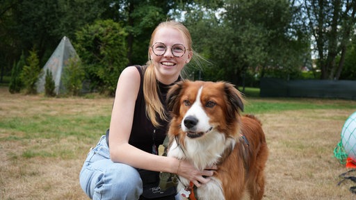 Eine Frau lächelt kniend in die Kamera und streichelt dabei einen Hund.