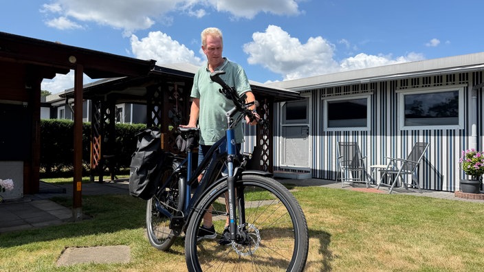 Ein Mann hält bei Sonnenschein im Garten eines Bungalows ein Fahrrad fest und schaut darauf.