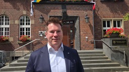 Ein Mann in Sakko und weißem Hemd, der vor einem Rathaus steht.