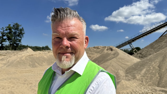 Jürgen Fröhlich, Pressesprecher von Holemans, steht vor dem Förderband vor Sand- und Kieshügeln und trägt eine grüne Warnweste.