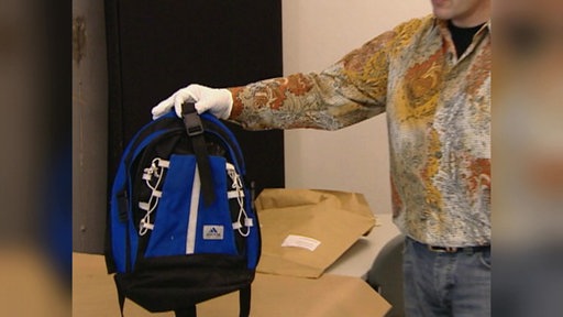 Ein blauer Adidas-Rucksack wird von einem Ermittler in der Hand gehalten.