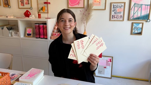 Künstlerin Sophia Wolf sitzt vor einer Wand mit bunten Bildern und hält Briefe in der Hand