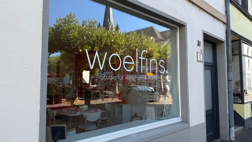Das Schaufenster des Ladens, in dem sich Bäume und eine Kirche spiegeln. Darauf steht in weißen Buchstaben "woelfins"