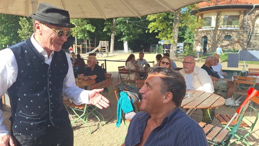 Mann mit Hut und Sonnenbrille redet mit einem Mann, der an einem Tisch sitzt