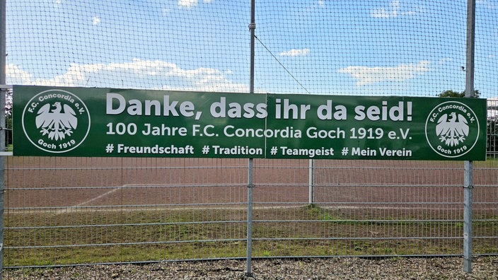 Grünes Banner des FC Concorida Goch hängt am Zaun des Fußballfeldes. Auf dem Banner steht "Danke, dass ihr da seid!" mit weißer Schrift.