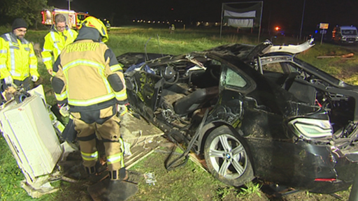 Drei Feuerwehrleute stehen neben einem durch einen Unfall zerstörten schwarzen Auto