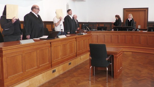 Angeklagte, ihre Verteidiger und Richter stehen im Gerichtssaal.