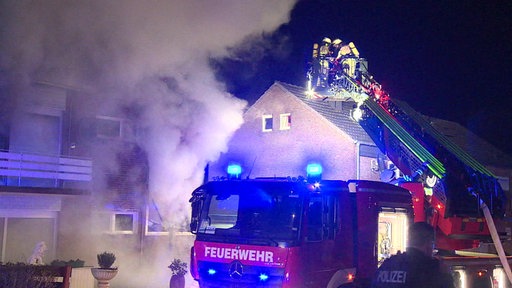 Ein Feuerwehrauto vor einem brennenden Haus. Die Drehleiter ist ausgefahren und mehrere Feuerwehrleute sind in dem Korb.