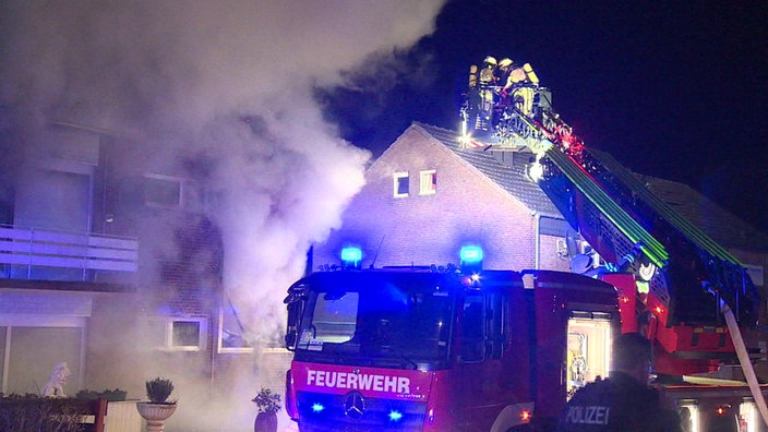 Ein Feuerwehrauto vor einem brennenden Haus. Die Drehleiter ist ausgefahren und mehrere Feuerwehrleute sind in dem Korb.