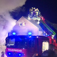 Ein Feuerwehrauto vor einem brennenden Haus. Die Drehleiter ist ausgefahren und mehrere Feuerwehrleute sind in dem Korb.