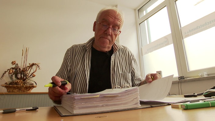 Peter Heß, ein älterer Mann mit Brille, sitzt in seinem Büro und markiert Dokumente in einem Aktenordner mit Textmarkern.