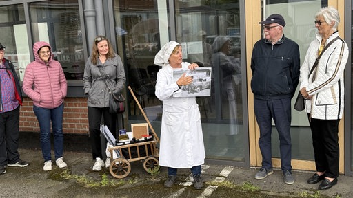 Eine Dame in einer Bäckeruniform hält ein Bild hoch und zeigt darauf, während sie weitere Personen anschauen.