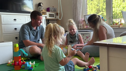 Mia sitzt mit ihrer Schwester und ihren Eltern auf einer grünen Matte im Zimmer und spielt.