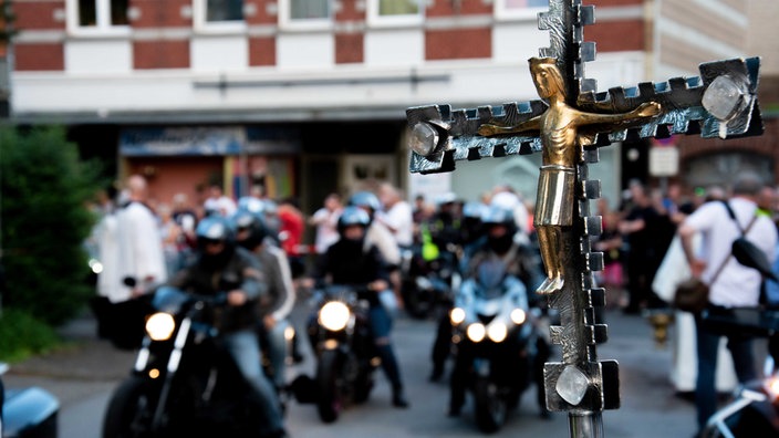 Kreuz mit Jesus-Statue im Vordergrund, unscharfe Motorräder im Hintergrund.