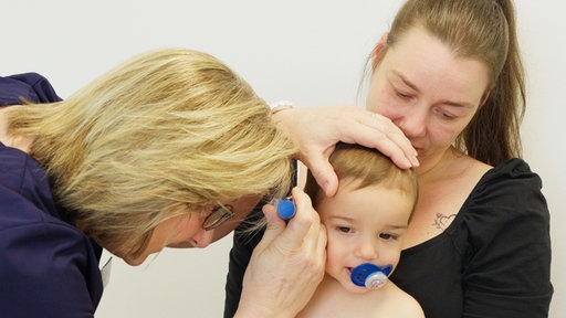 Kinderärztin untersucht Ohren eines Kleinkinds, das auf dem Schoß seiner Mutter sitzt