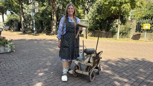 Eine Frau in historischer Kleidung lächelt in die Kamera. Neben ihr ein Bollerwagen mit Utensilien.
