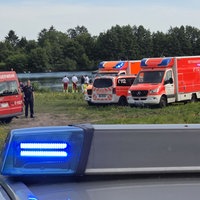 Im Hintergrund ist ein See, davor stehen einige Menschen in Rettungs-Uniformen. Rechts daneben stehen mehrere Krankenwagen und ein Notarztwagen. Links im Bild sind noch mehr Rettungsfahrzeuge. Im Vordergrund sieht man das Dach eines Polizeiautos.