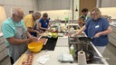 Um eine Kücheninsel herum stehen fünf Personen und kochen.