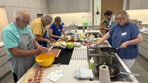 Um eine Kücheninsel herum stehen fünf Personen und kochen.