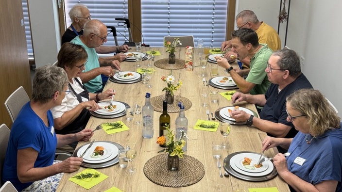 An einem großen Holztisch sitzen insgsamt acht ältere Personen und essen gemeinsam. 