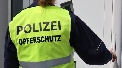 Eine Person von hinten, die an einer Tür klingelt. Über der Polizeiuniform trägt sie eine neongelbe Weste mit der Aufschrift "Polizei Opferschutz".