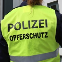 Eine Person von hinten, die an einer Tür klingelt. Über der Polizeiuniform trägt sie eine neongelbe Weste mit der Aufschrift "Polizei Opferschutz".