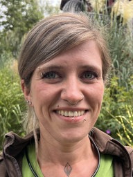Lara Schmitt (Tierpflegerin im Tiergarten Kleve) steht draußen im Grünen, trägt ein grünes Shirt mit einer braunen Jacke und grinst.