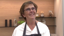 Chefköchin Monika Mott, eine Frau mit Brille und Kochschürze, lächelt in die Kamera