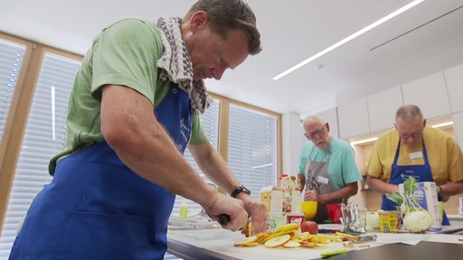 Ein Mann mit Kochschürze schneidet einen Apfel in Scheiben. Im Hintergrund stehen zwei weitere Männer an einer Kochinsel und kochen.
