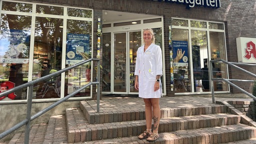 Tatjana Kremer trägt einen weißen Kittel und hat schulterlanges, blondes Haar. Sie steht vor der Apotheke am Stadtgarten auf einer Treppe.